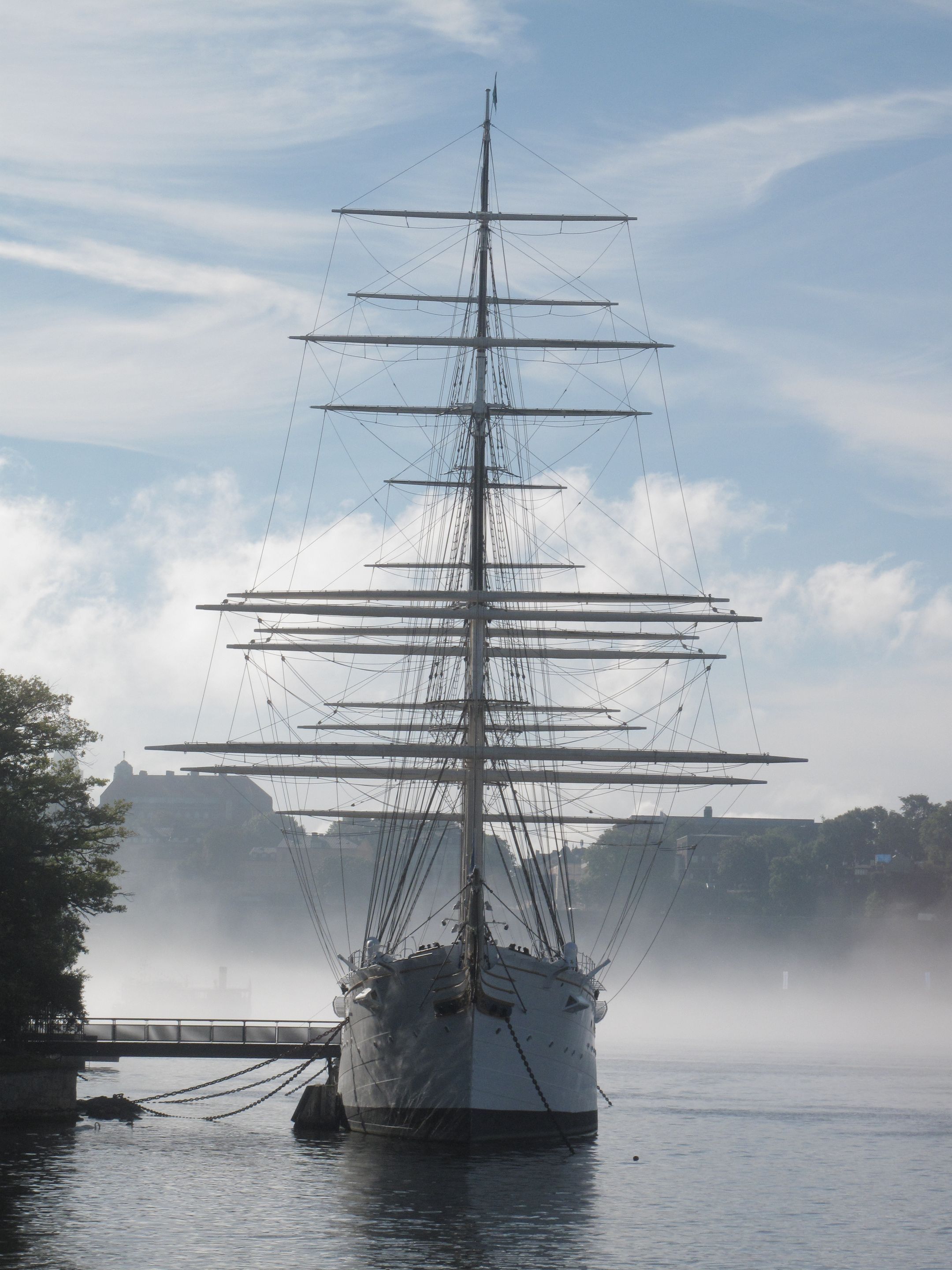 Skeppet af Chapman ligger förankrad vid Skeppsholmen västra strand, fotograferad rätt framifrån från Skeppsbron. I bakgrunden syns morgondimman och bortom den Södermalms höjder. Himmlen bakom skeppets master är blå men molning.